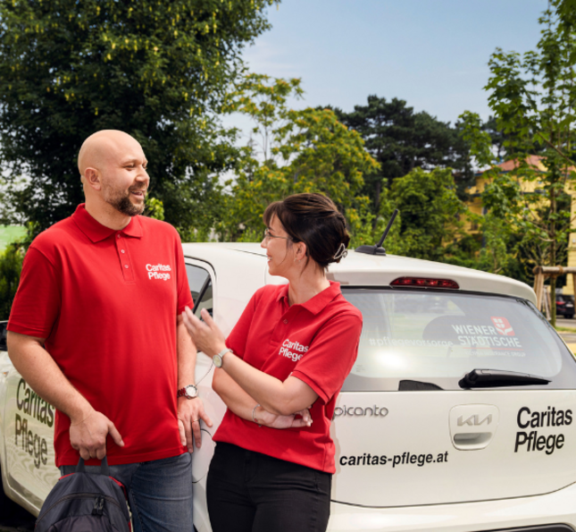 Caritas Pflege - auch mobil unterwegs Zwei Mitarbeiter*innen der Caritas Pflege in roten Caritas T-Shirts unterhalten sich vor ihrem Caritas Pflege-Auto