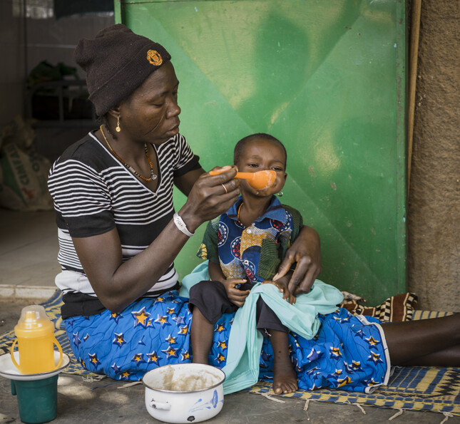 Viele Kindern in Burkina Faso leiden an Unterernährung - die Caritas hilft. © Jork Weismann Frau sitzt mit Kleinkind auf dem Schoß und füttert es mit Löffel
