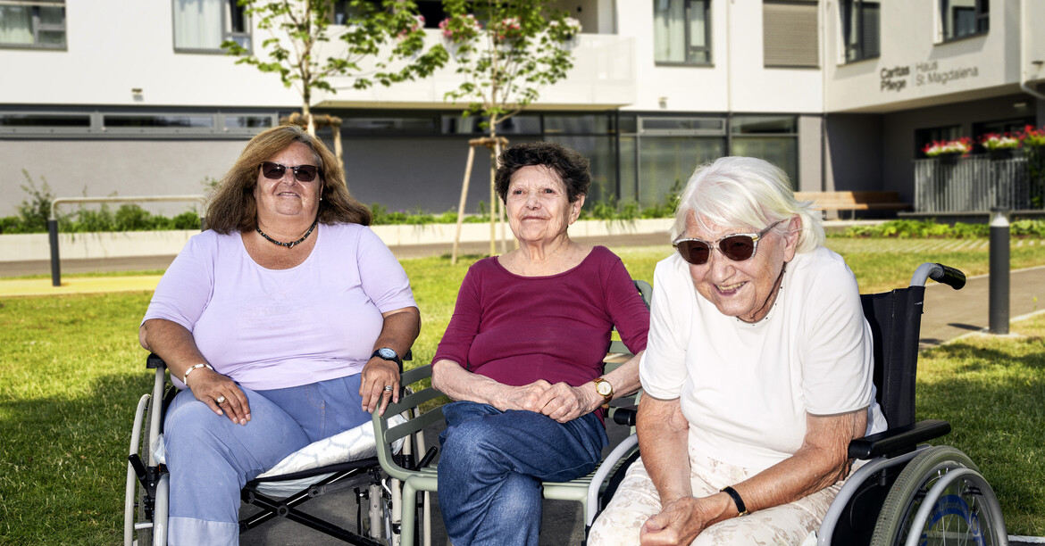 Caritas Pflege - wenn Omis zu Homies werden. © Norbert Kniat Drei ältere Frauen sitzen auf ihren Rollatoren bzw. Rollstühlen nebeneinander, sie lächeln in die Kamera. Im Hintergrund ist ein Pflegewohnhaus zu erkennen.