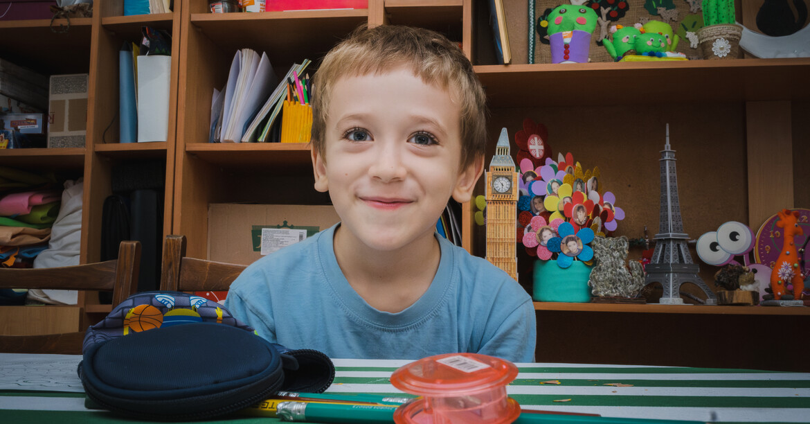 Junger Schüler sitzt lächelnd mit Stiften und Büchern an Schultisch
