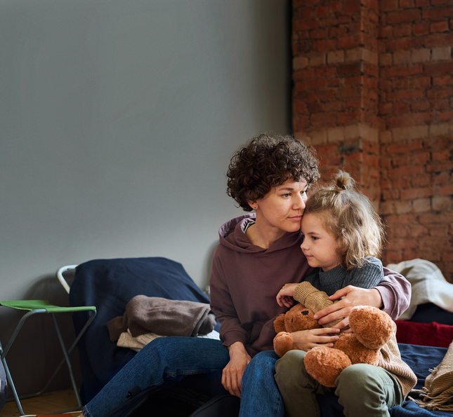 Eine Frau sitzt auf einer Bank, auf ihrem Schoß ein kleines Kind mit Kuscheltier in der Hand.