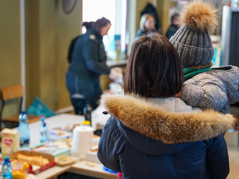 Eine Frau in Winterkleidung, die ein Kind auf dem Arm hat, steht vor einer Sachspendenausgabe.
