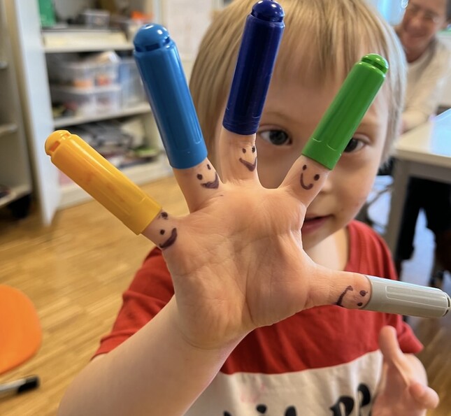 Kleinkind zeigt bemalte Finger mit Stiftkappen als Figuren in Betreuungsraum